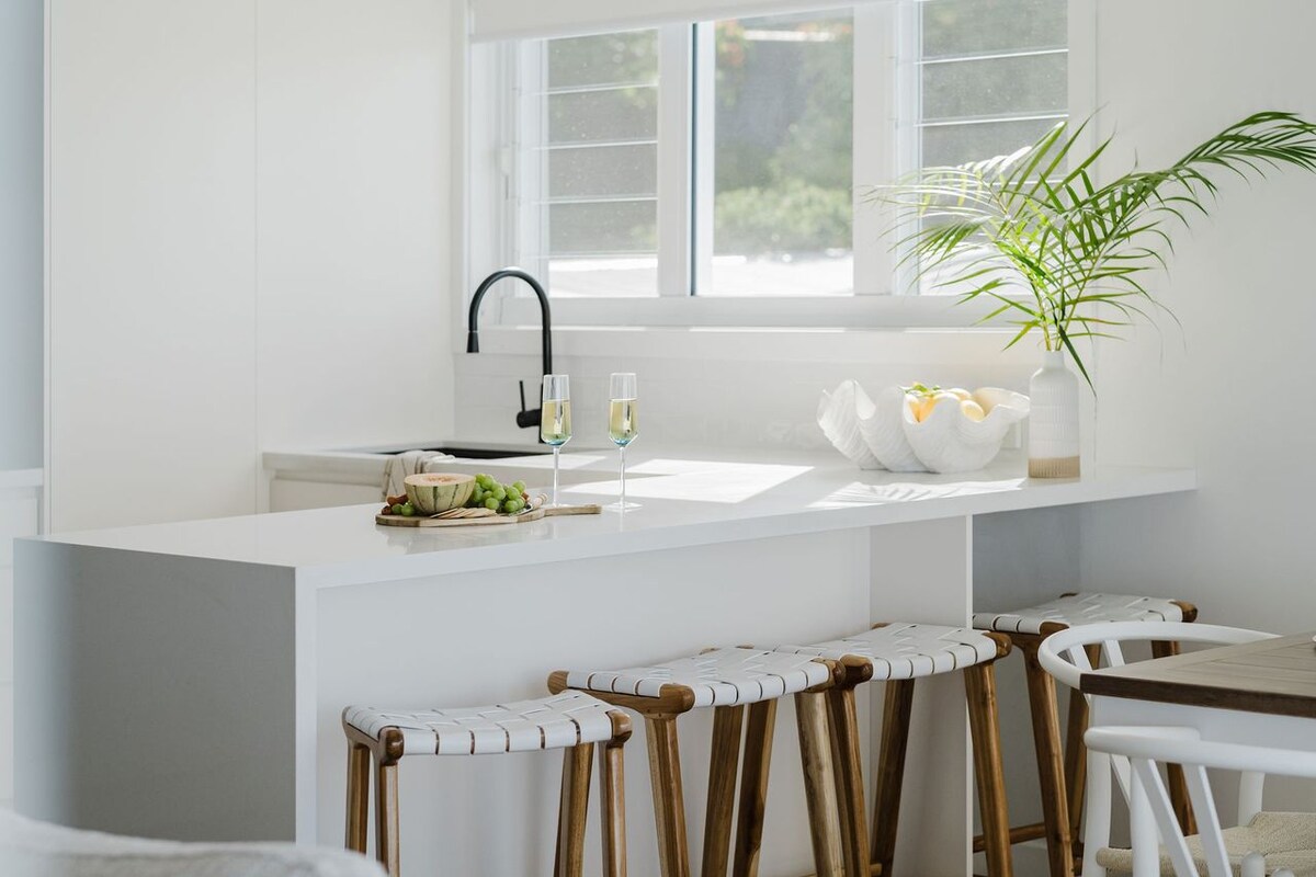 A clean and modern kitchen space features white cabinetry and a sleek countertop. Four wooden stools with white textile seating line a breakfast bar. Fresh greenery and a bowl of fruit are displayed on the counter. Natural light fills the area through large windows.