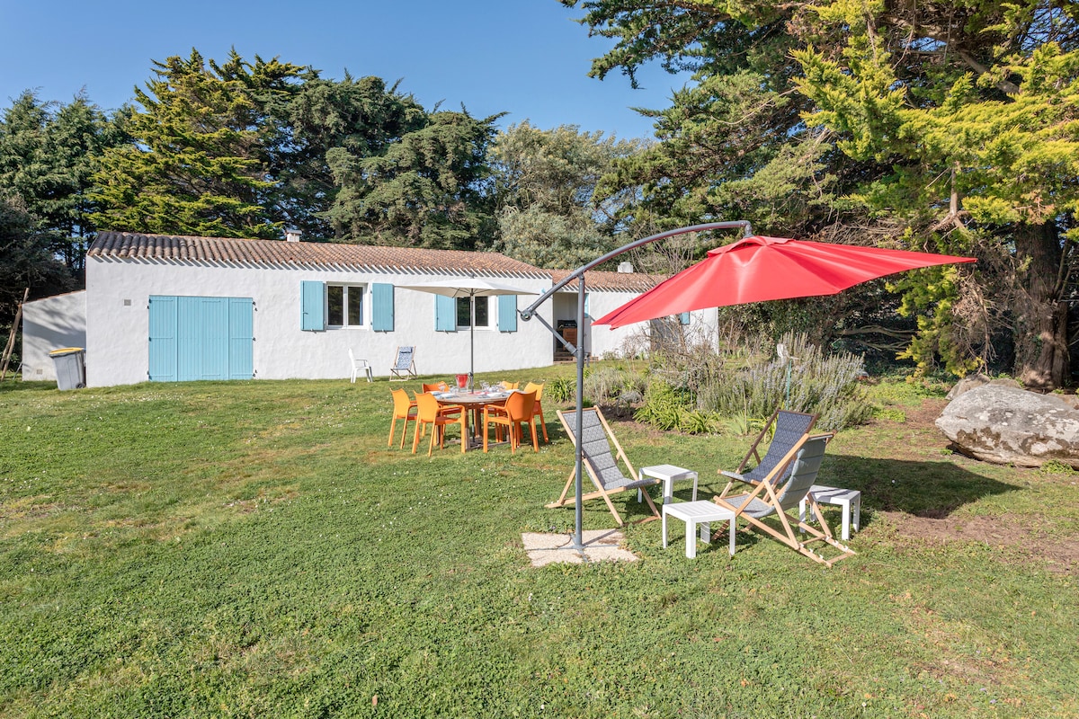 The exterior of the holiday home is surrounded by a spacious garden, featuring a red umbrella-shaded seating area with a table and colorful chairs. Two lounge chairs are positioned nearby for relaxation, framed by lush greenery and trees that provide privacy to the space.