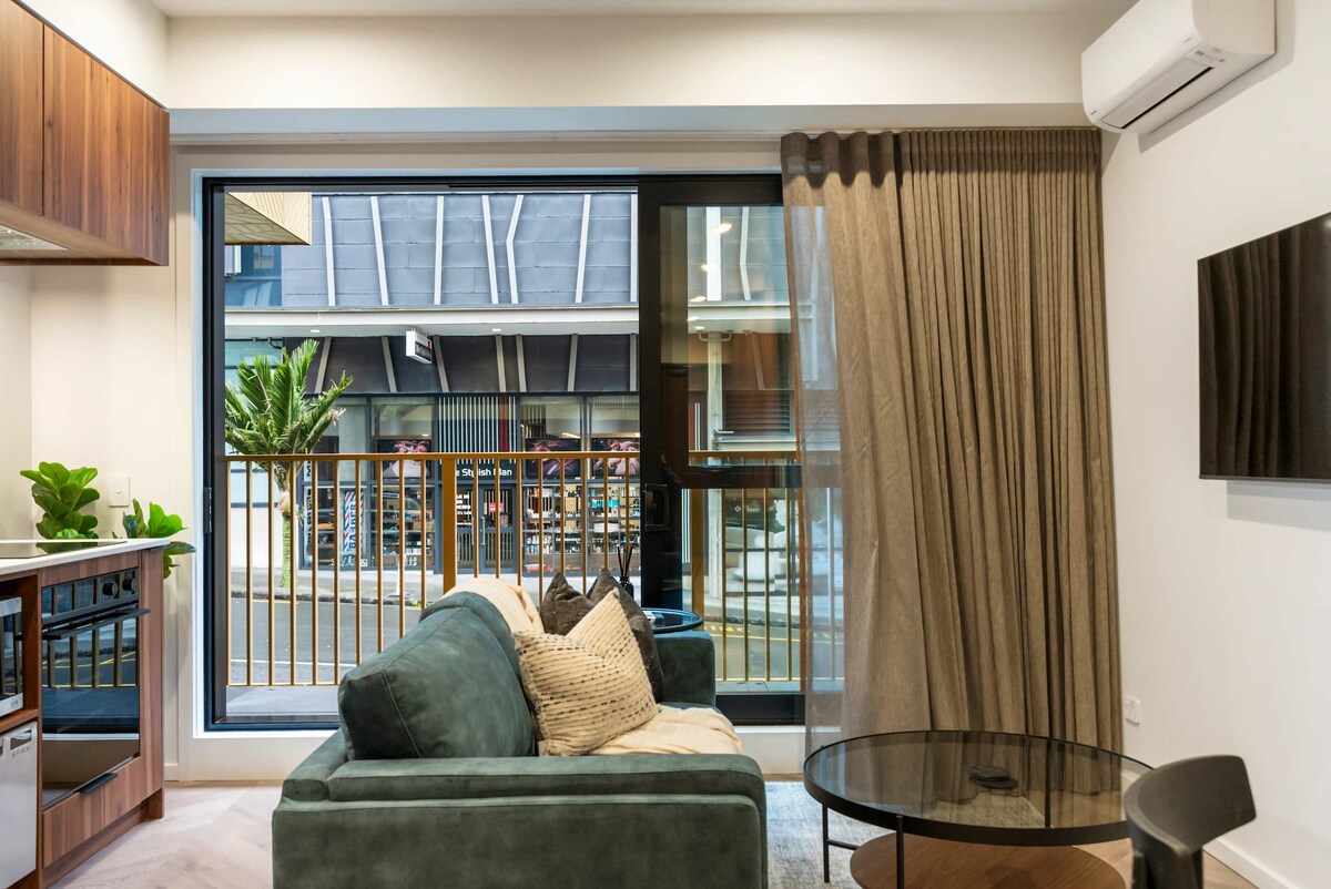 A comfortable living area features a plush sofa, adorned with decorative cushions. A round glass table sits nearby, complemented by a view of the balcony through large windows. Natural light filters gently through sheer curtains, enhancing the inviting ambiance of the space.