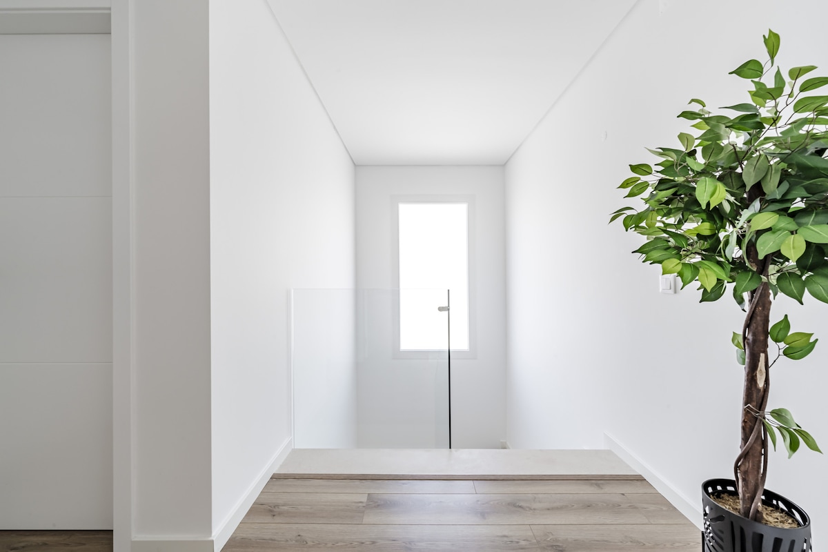 A well-lit hallway presents a minimalist design with white walls and warm wooden flooring. A large window permits natural light to fill the space, complementing the contemporary feel. A decorative potted plant adds a touch of greenery, enhancing the serene ambiance.