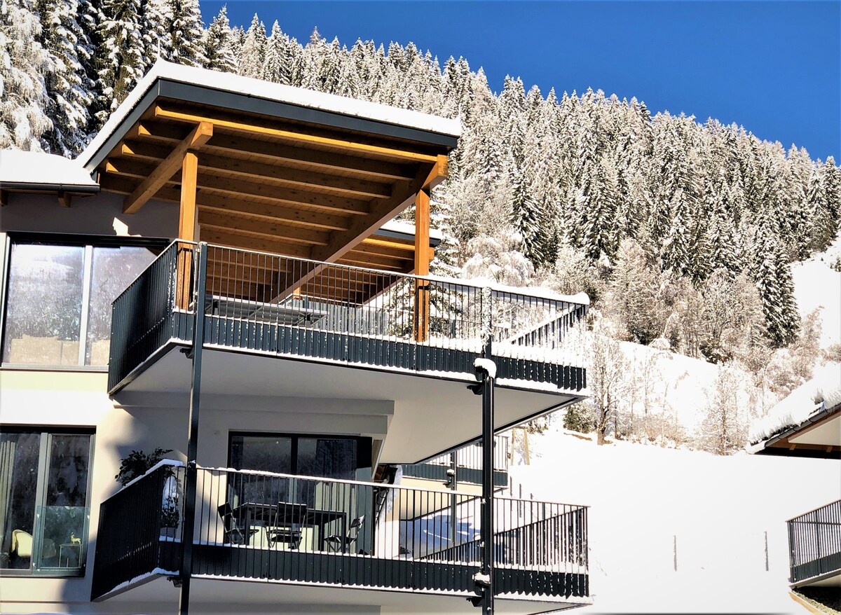 A modern flat is showcased with a wooden-covered balcony situated under a clear blue sky. Surrounding snow-covered mountain trees provide a serene backdrop, while the balcony features outdoor seating, inviting guests to enjoy the winter landscape.