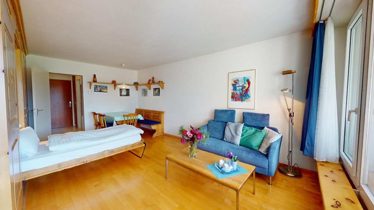 A well-furnished living area features a fold-away bed, a blue sofa, and a wooden dining table with chairs. A bright artwork adorns the wall, and fresh flowers are placed on a coffee table. Large windows allow natural light to illuminate the space.