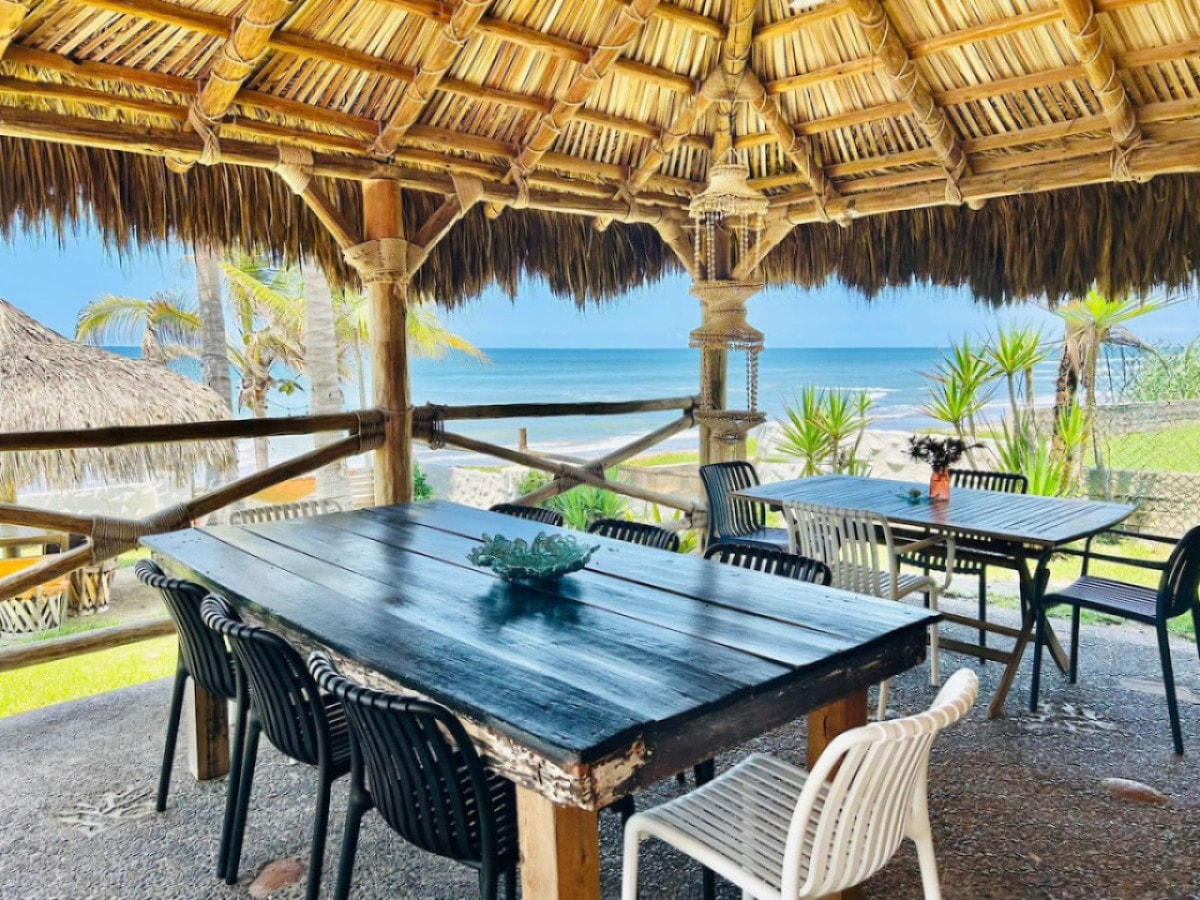 An outdoor dining area is presented under a thatched roof with a wooden table surrounded by black and white chairs. Lush greenery and palm trees are visible in the background, along with a clear view of the ocean, creating a serene setting for meals.