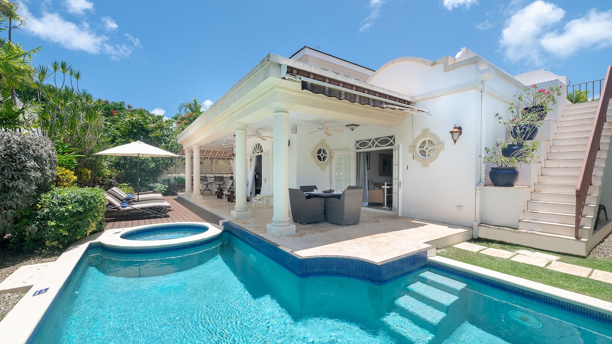 A private pool area features a clear blue pool and a shaded gazebo, perfect for outdoor dining. The villa's white façade is complemented by tropical greenery. Lounge chairs are placed beside the pool, with stairs leading to the upper level of the home.