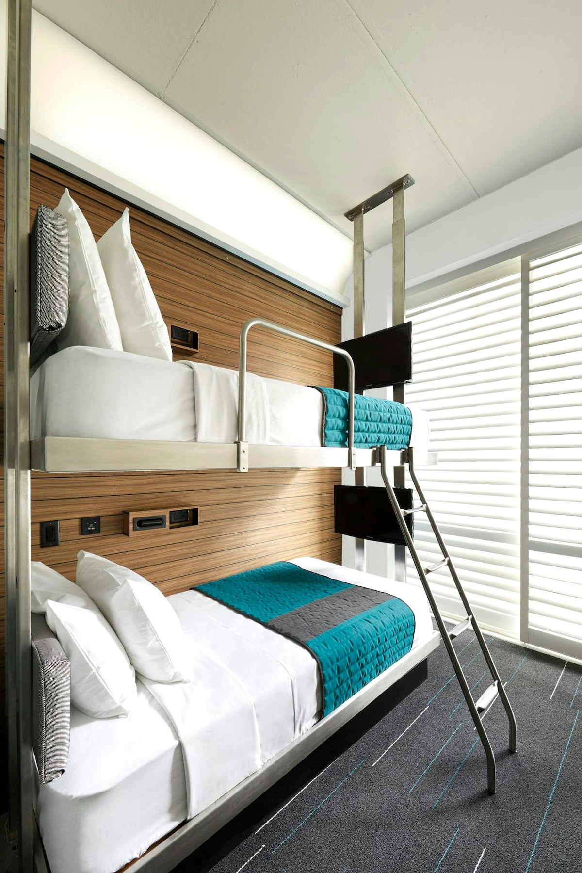 A compact guest room features a bunk pod design, with a spacious bottom bed and a raised top bed. Both beds are dressed in crisp white linens with turquoise accents. The room includes a large window with adjustable shutters, allowing natural light to filter in.