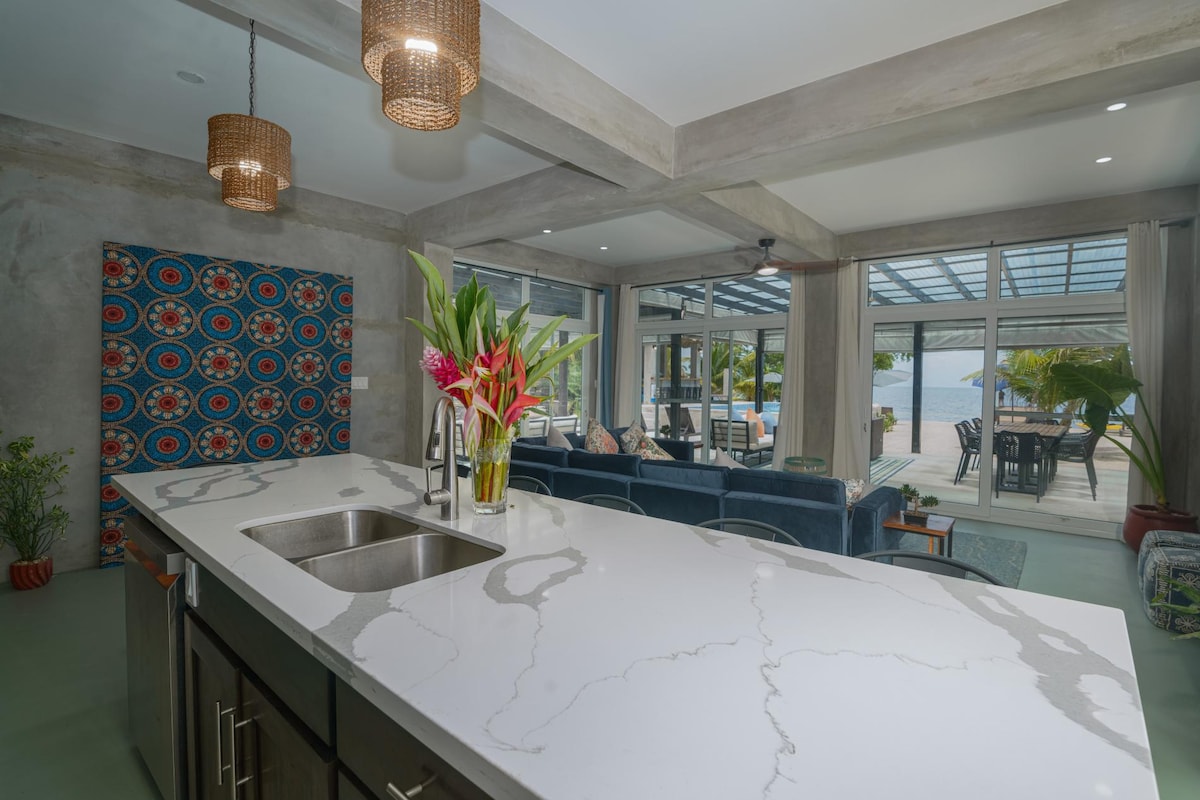 A modern kitchen is showcased with a sleek countertop featuring prominent veining. Fresh flowers are arranged in a vase, adding a lively touch. The open layout allows visibility to a spacious living area beyond, with large windows providing a view of the outdoors and the Caribbean Sea.