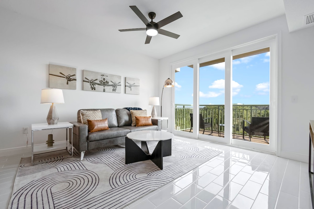 The modern living room features a spacious sofa complemented by decorative pillows, along with a sleek coffee table atop an area rug. Large glass doors open to a private balcony, revealing expansive views. Ceiling fan and lamps provide ambient lighting, enhancing the room's inviting layout.