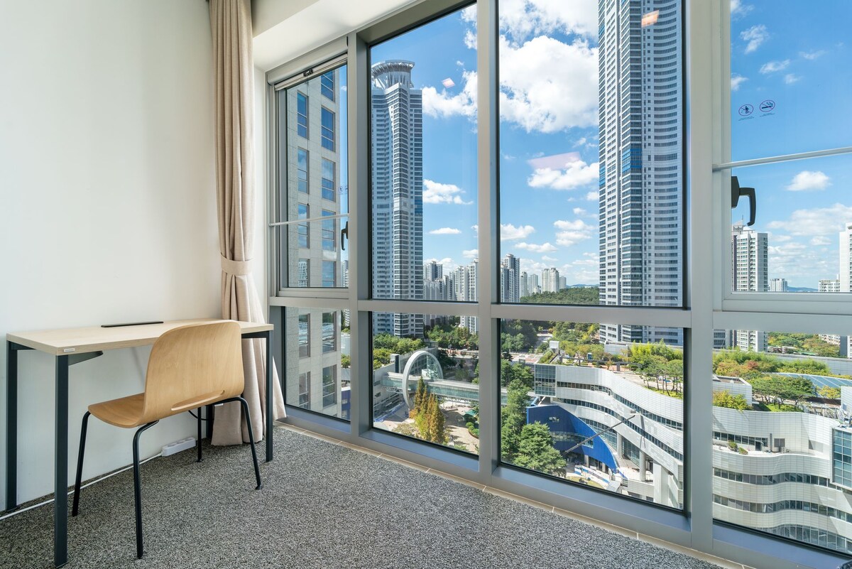 A bright room features a simple wooden desk and chair positioned near a large window. Cityscape views highlight contemporary buildings and greenery outside, with natural light enhancing the space. The carpeted floor adds comfort to the overall atmosphere.