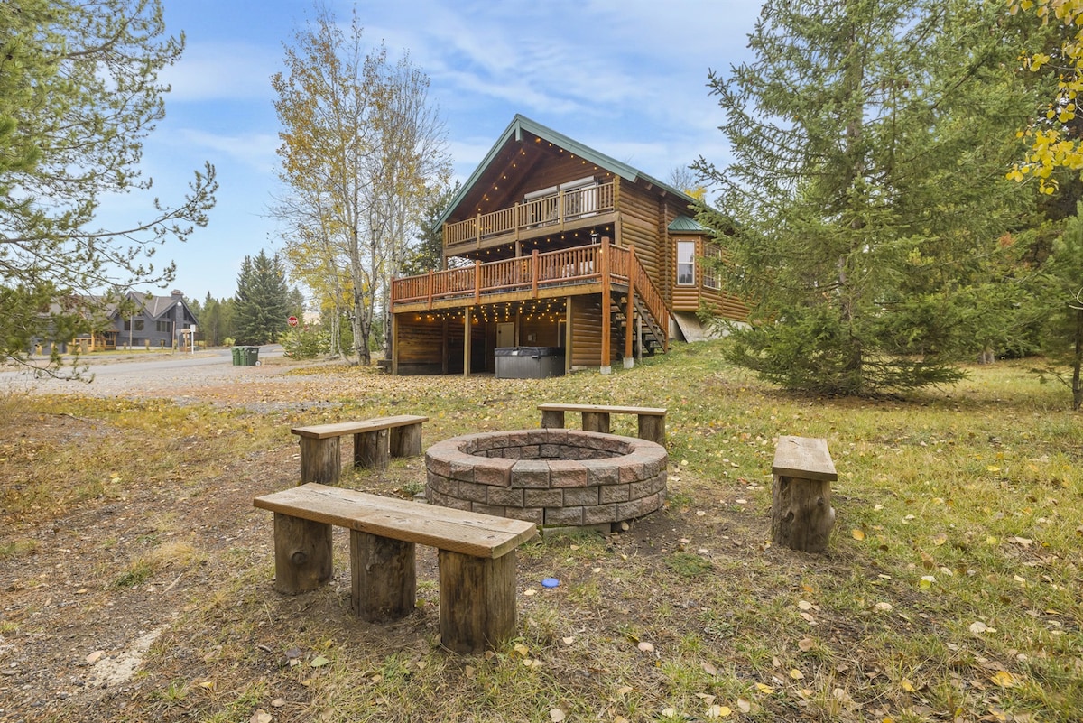 A constructed fire pit is surrounded by rustic wooden benches, situated in a spacious outdoor area with scattered trees. The modern cabin, with a green roof and wooden exterior, sits prominently in the background, offering access to outdoor gatherings.