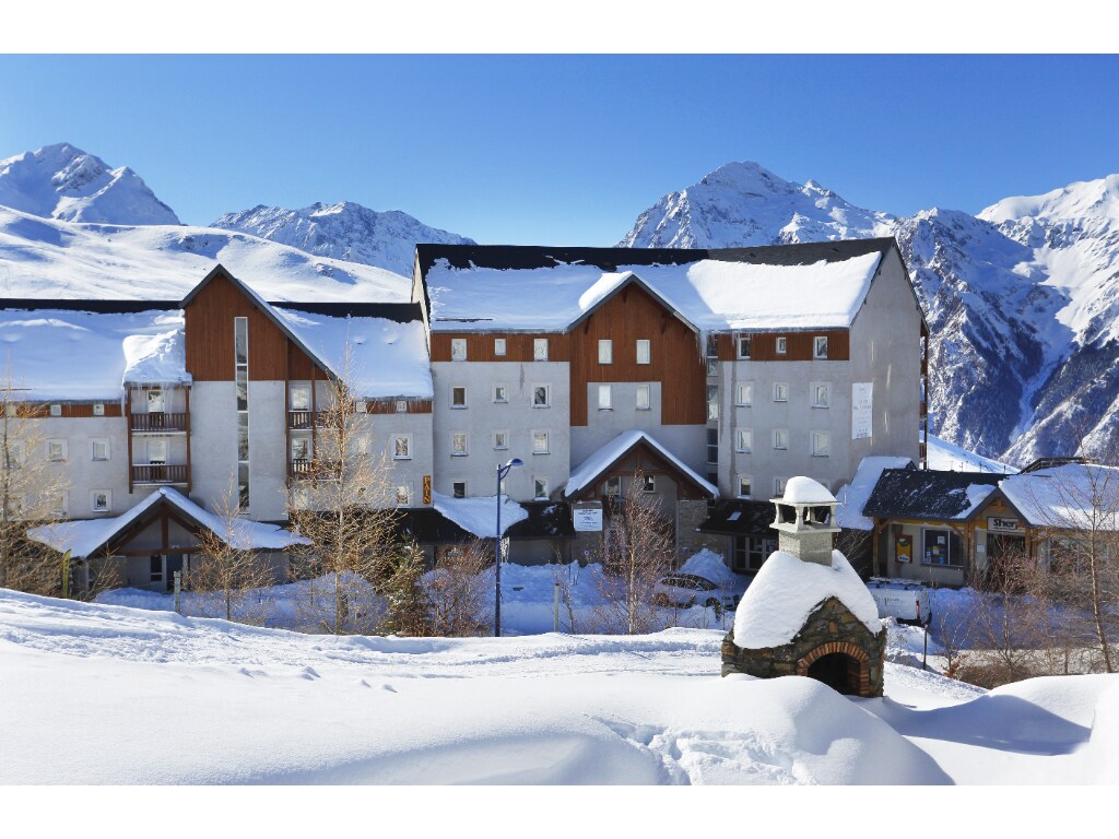 The exterior of the residence is showcased amidst a winter landscape, with snow blanketing the ground. The building features a combination of wood and stone facades, complemented by balconies and a chimney. Majestic mountain peaks are visible in the background under a clear blue sky.