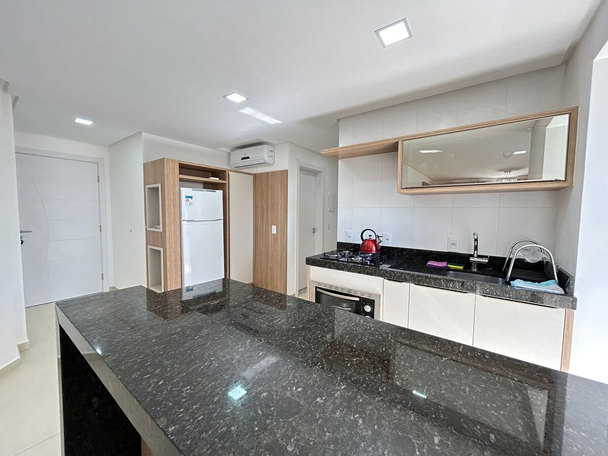 The kitchen area features modern cabinetry and a dark stone countertop, complemented by light-colored walls and a large mirror. A refrigerator is positioned near the entrance, while a kettle and sink are visible on the counter, enhancing functionality and convenience.