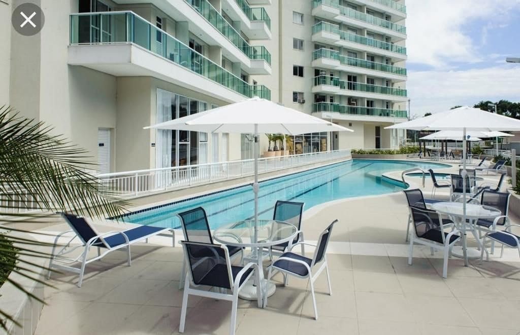 An inviting outdoor area features a swimming pool bordered by stylish lounge chairs and tables with umbrellas for shade. The building's modern architecture provides a contemporary backdrop, and the pool area is enhanced by well-maintained surrounding greenery.