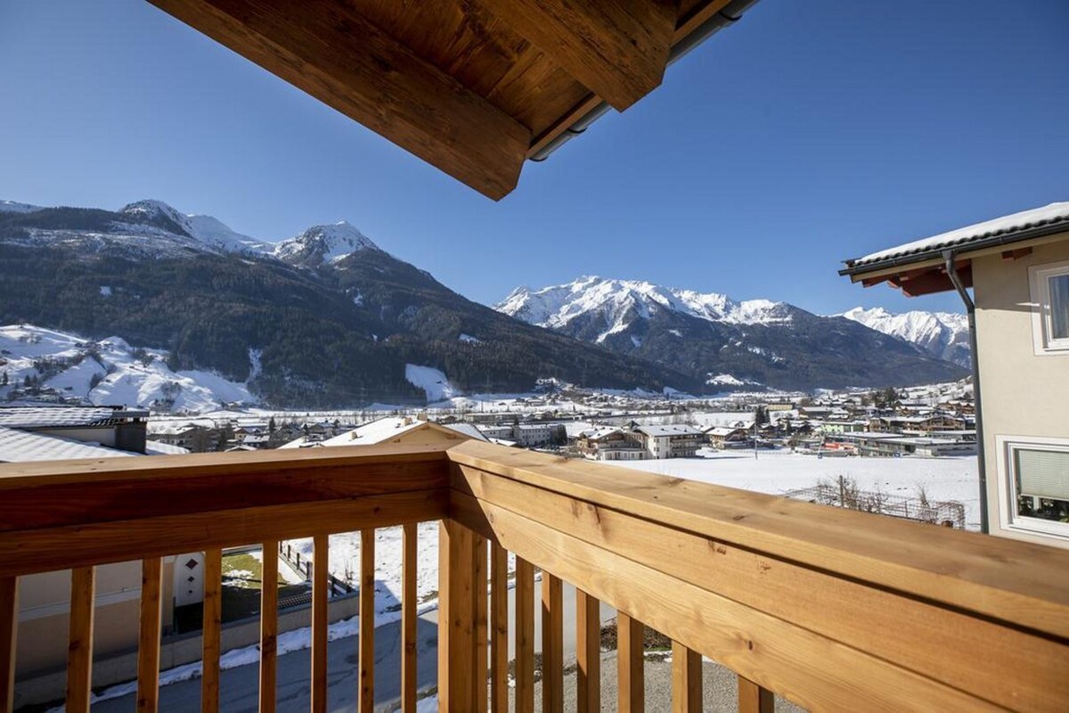 A wooden balcony railing frames a stunning view of snow-covered mountains under a clear blue sky. The expansive valley below is dotted with charming buildings, creating a serene winter landscape.