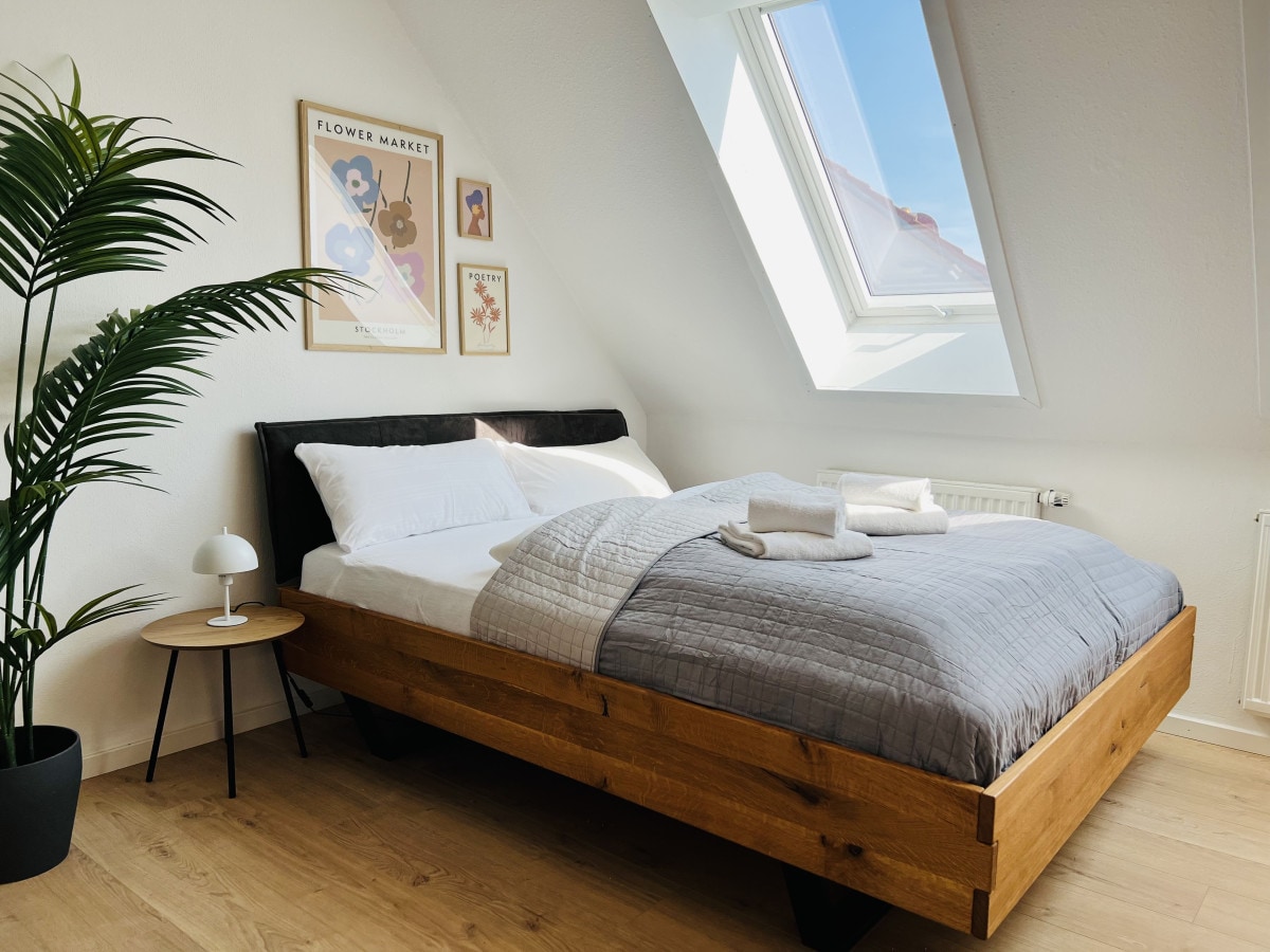 A cozy sleeping area features a wooden bed with soft linens and neatly arranged towels. A modern bedside table holds a lamp, and a framed artwork adorns the wall above. Natural light pours in through the slanted skylight, illuminating the calm, minimalist decor.