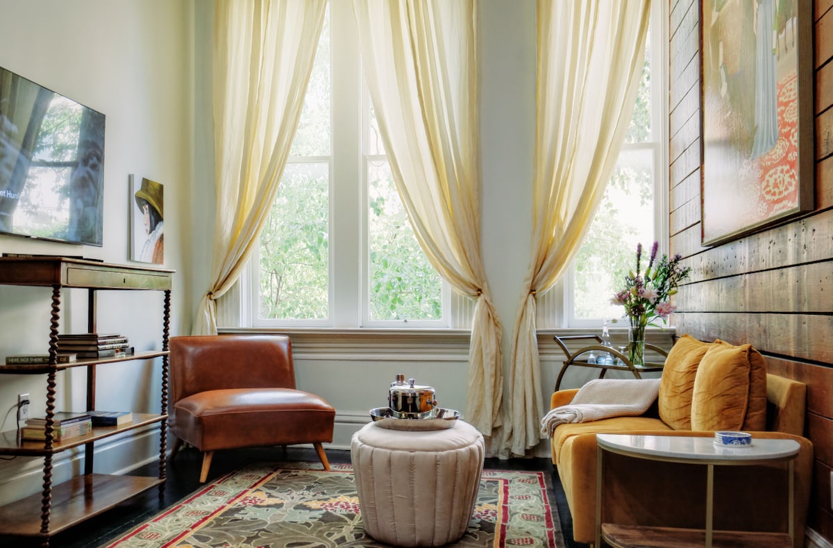 A cozy sitting area features large windows dressed with light curtains, allowing natural light to fill the space. A stylish brown chair and yellow accent chair are positioned beside a decorative pouf and a small side table. A floral arrangement adds a touch of color to the room.