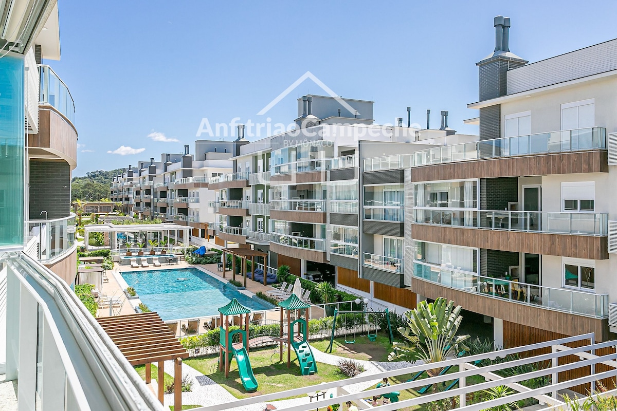 A view of the condominium grounds reveals multiple modern buildings arranged around a central pool area. The pool is surrounded by lounge chairs, and a children's play area with colorful equipment is visible in the foreground, framed by lush landscaping.
