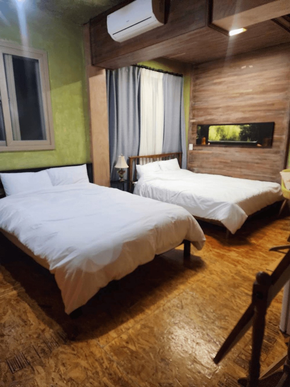 A room is presented with two spacious beds, each featuring white linens. Natural light enters through a window covered with sheer curtains. A wooden accent wall adds warmth, complemented by a decorative shelf displaying greenery. A bedside lamp is positioned next to one of the beds.