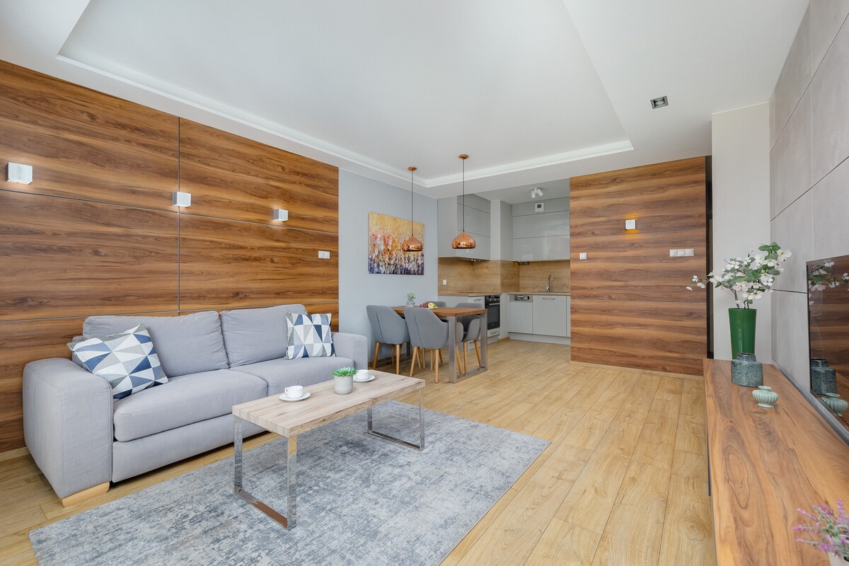 Светла всекидневна с удобен разтегателен диван и модерни дървени акценти. Помещението включва мек килим и зона за гледане на телевизия, идеална за спокойни вечери.