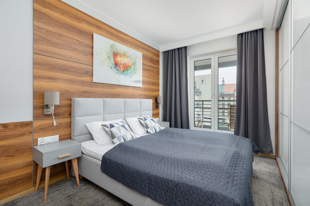 Спокойна спалня с двойно легло, украсено със сиво спално бельо. Дървените панели зад леглото създават топла и модерна атмосфера.
