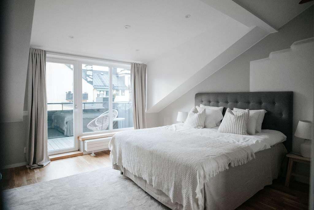 The bedroom features a spacious king-sized bed dressed in soft, neutral linens. Large glass doors provide access to a private balcony, bathing the room in natural light. Soft curtains frame the windows, complementing the minimalist decor in a calming color palette.