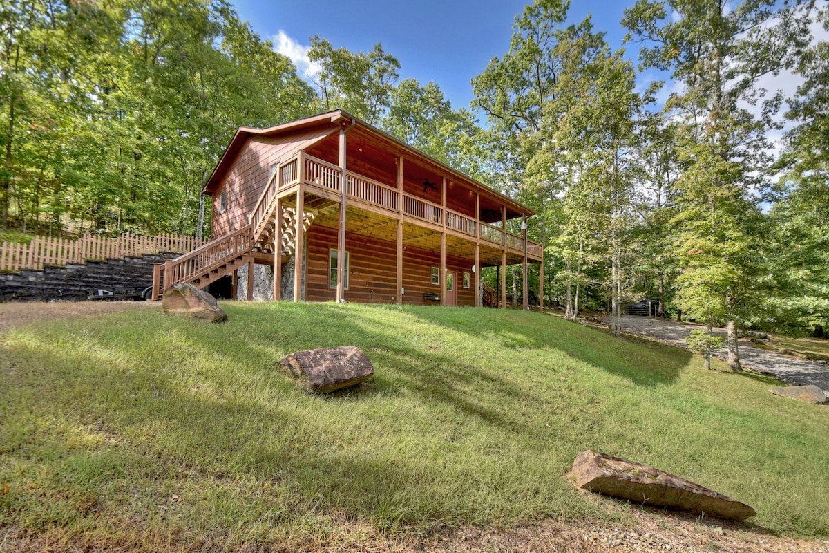 The exterior of a rustic cabin is framed by lush greenery, showcasing wooden siding and a wrap-around porch. Large steps lead to the entry, with an inviting landscape featuring natural stone and grassy slopes. Sunlight casts gentle shadows on the building's surface.