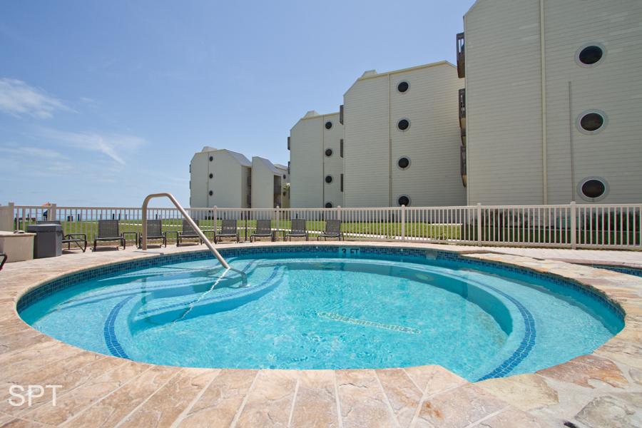 A circular hot tub is positioned near a resort building, featuring clear blue water that reflects the sunlight. A white railing surrounds the area, ensuring safety. The backdrop consists of multiple light-colored structures, enhancing the serene poolside atmosphere.