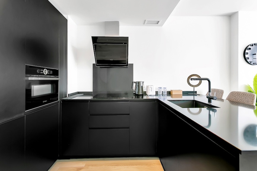The modern kitchen features sleek black cabinetry and appliances, including a built-in oven and a cooktop. A minimalist layout is enhanced by a large black countertop and a stylish faucet. Natural light streams in, highlighting the contemporary design.