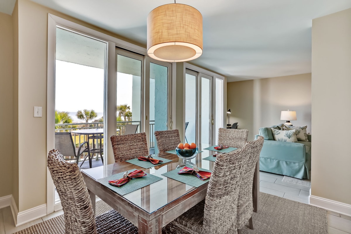 A dining area is showcased with a wood table and six woven chairs, set with colorful placemats and napkins. Natural light floods through sliding glass doors, offering a view of palm trees and the balcony. A round pendant light hangs above, adding a modern touch.