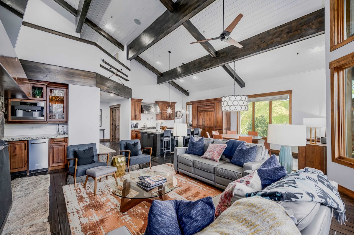 An open-concept living area showcases exposed wooden beams and large windows that invite natural light. Cozy seating arrangements include plush couches and armchairs set around a coffee table, while a patterned area rug adds warmth to the space. The kitchen features modern appliances and stylish cabinetry.