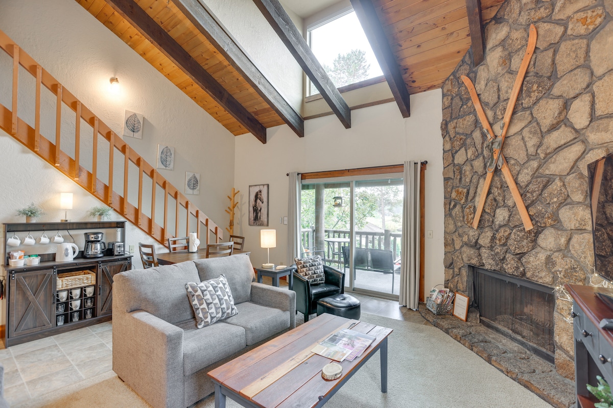 A spacious living area features vaulted ceilings and natural wood beams. A cozy grey sofa and black seating are arranged around a wooden coffee table. Large sliding glass doors lead to a private balcony, while a stone fireplace adds warmth to the room.