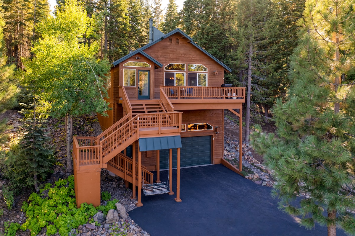 The exterior of a spacious wooden home is showcased amidst a natural setting, featuring large windows that allow for ample sunlight. A staircase wraps around the side, leading to a welcoming deck area. Surrounding pine trees enhance the serene atmosphere.