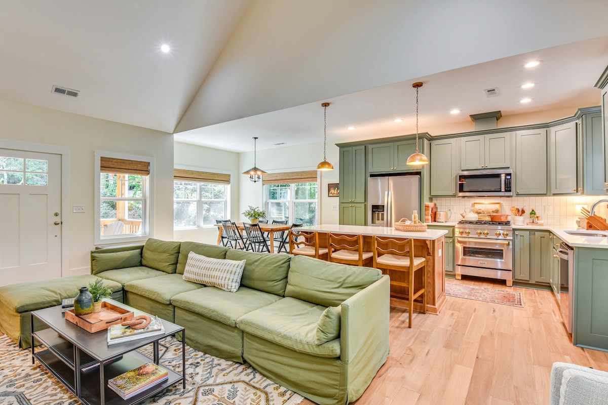 A spacious living area is highlighted by a light green sectional sofa, complemented by a patterned area rug. A kitchen with green cabinetry and stainless steel appliances is visible in the background, alongside a dining table with multiple chairs, creating a welcoming communal space.