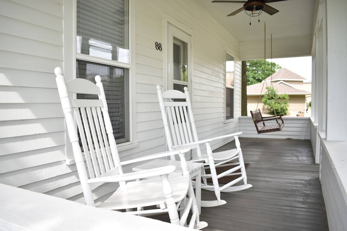 A spacious front porch features two white rocking chairs and a swing, providing comfortable seating. Natural light filters through the windows, enhancing the relaxing atmosphere. The porch extends along the house, with a smooth wooden floor adding to its inviting appeal.