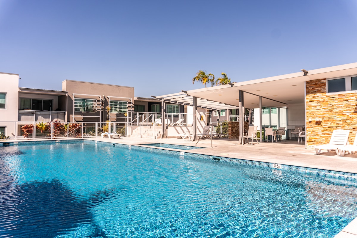 A large swimming pool reflects sunlight under a clear blue sky, surrounded by modern buildings. Lounge chairs are positioned around the pool area, and a shaded patio is visible in the background, offering additional seating options.