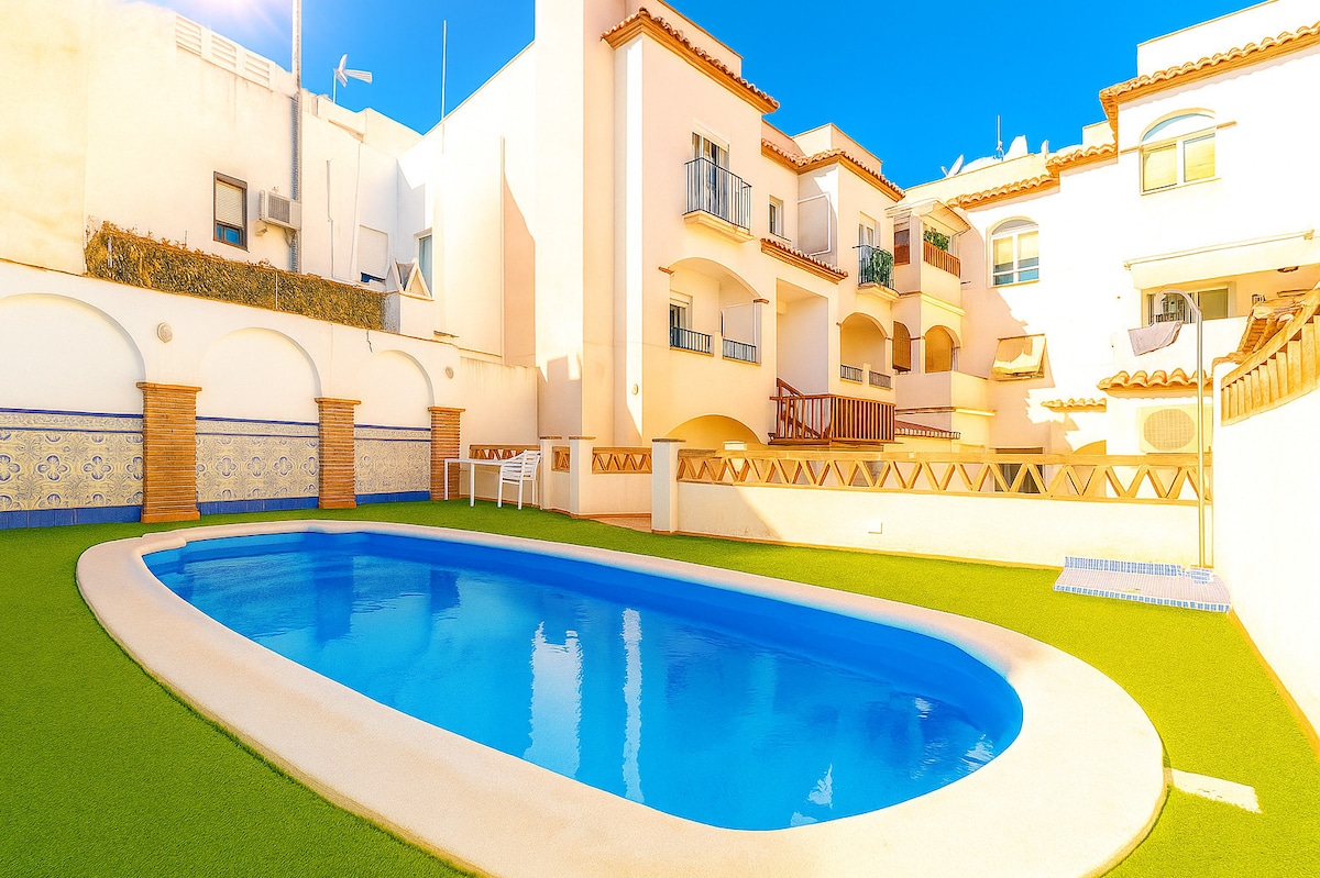 A communal swimming pool is surrounded by well-maintained grass and white walls. The pool's clear water reflects the sunlight, while nearby balconies overlook the area. Sun loungers can be spotted on the deck, offering a place for relaxation.