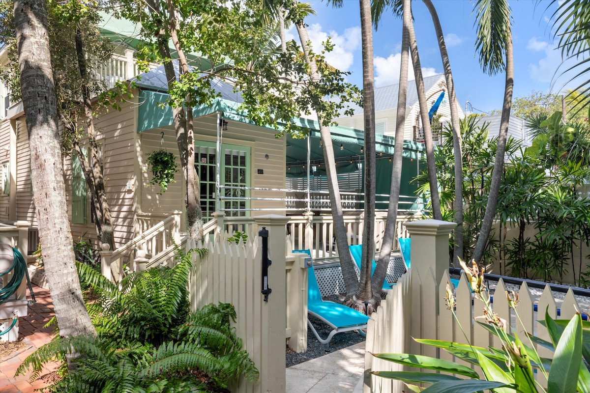 The exterior of the historic condo is framed by lush greenery and tall palm trees, creating a serene atmosphere. Loungers in vibrant blue are positioned on the wraparound porch, inviting relaxation. The entrance is bordered by a charming white picket fence, enhancing the inviting environment.