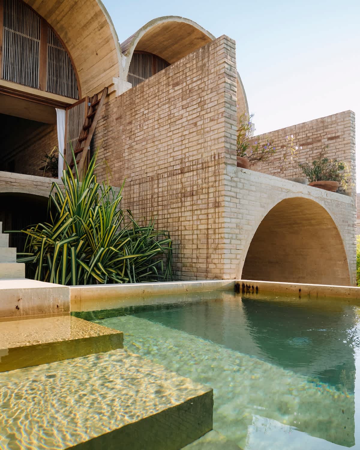 A serene view of a private pool with tranquil water, reflecting the natural surroundings. A unique architectural structure with arched elements rises in the background, complemented by greenery along the pool's edge. Steps lead out of the water, enhancing the connection to the space.
