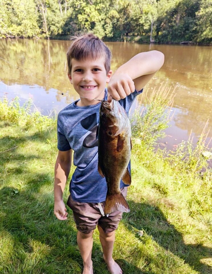 A child stands on grassy ground next to the Muskegon River, holding a freshly caught smallmouth bass. The calm water reflects the surrounding greenery, creating a serene backdrop for this outdoor fishing experience.