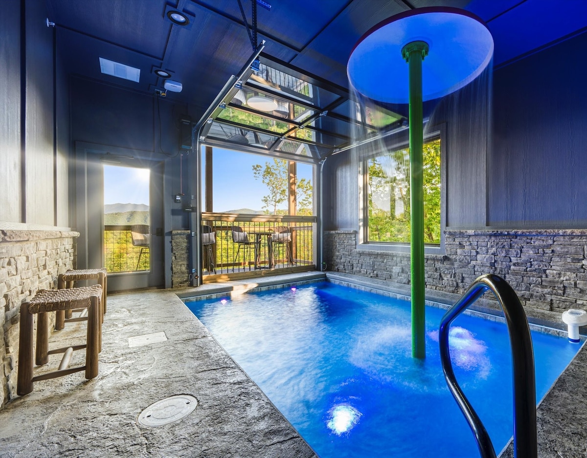 An indoor heated saltwater pool is visible, featuring a retractable garage door that opens to stunning mountain views. A large waterfall umbrella adds a unique touch while seating is situated nearby. Stone accents enhance the luxurious atmosphere of this relaxing space.