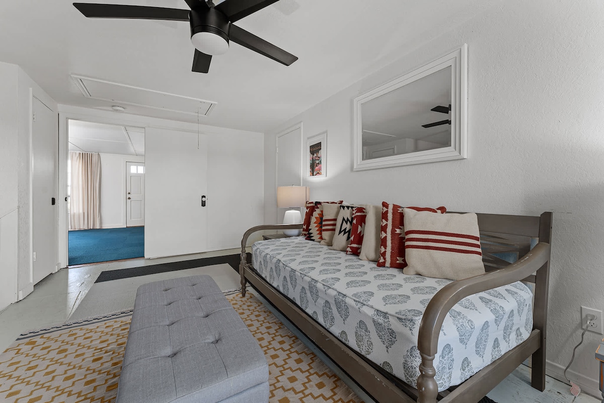 A daybed is positioned in a bright room, adorned with red and white decorative pillows and a patterned cover. A modern ceiling fan is installed, and light filters in through a nearby window. A neutral area rug and a small ottoman complete the inviting atmosphere.
