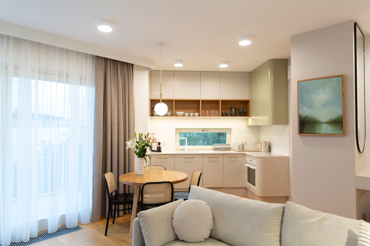 A well-lit open space features a modern kitchen with light cabinetry and a round dining table, surrounded by black chairs. Large windows draped with sheer curtains provide natural light, highlighting the soft textures of a gray sofa in the foreground.