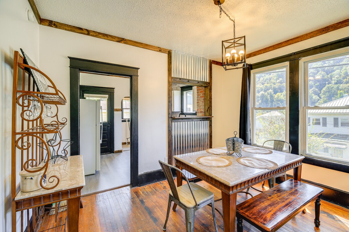 A dining area features a wooden table surrounded by four chairs and a bench. Natural light enters through two large windows, illuminating the space. A decorative metal shelf adds a unique touch, and a door leads to the well-equipped kitchen.