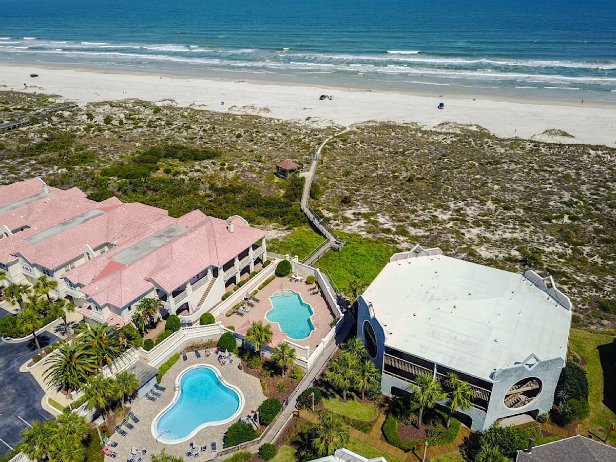 An aerial view captures the seaside condo complex, featuring inviting swimming pools surrounded by lush landscaping. The coastline stretches in the background, with soft waves lapping against the shore. A private boardwalk leads directly to the beach, enhancing accessibility to the outdoor environment.