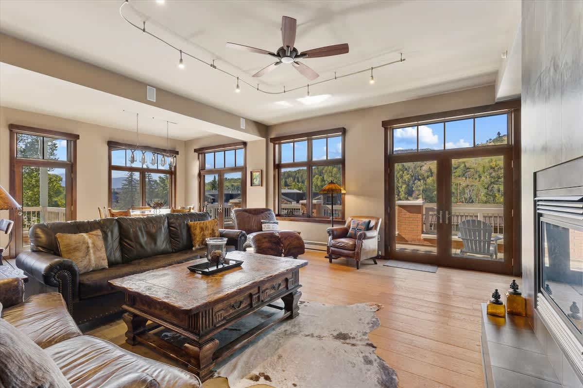 A spacious living area features a large brown leather sofa and a rustic wooden coffee table, complemented by a cozy gas fireplace. Sunlight floods through multiple windows, showcasing mountain views and inviting warmth. A dining area with a wooden table is visible in the background.