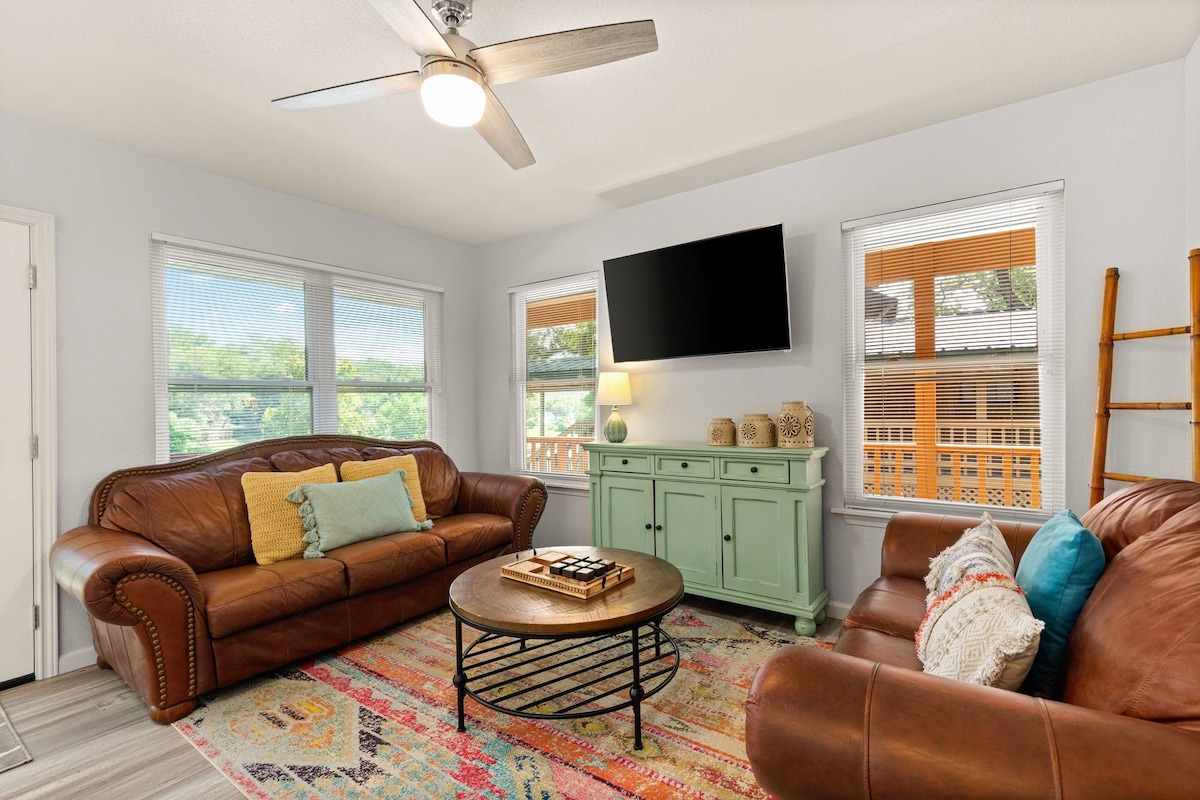 A welcoming living area is featured with two leather sofas placed around a round coffee table. A large flat-screen TV is mounted on the wall, and natural light fills the space through multiple windows adorned with blinds. A colorful area rug adds a touch of warmth.