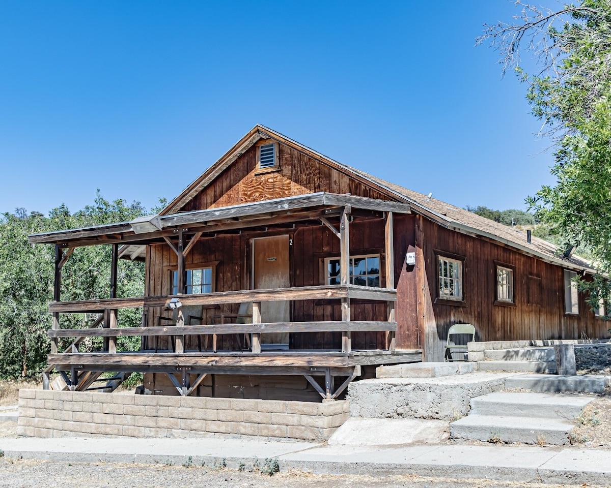 The rustic cabin exterior features warm wood tones and a covered porch with railing. Steps lead up to the entrance, surrounded by natural greenery. The building is set against a clear blue sky, showcasing a tranquil rural atmosphere.