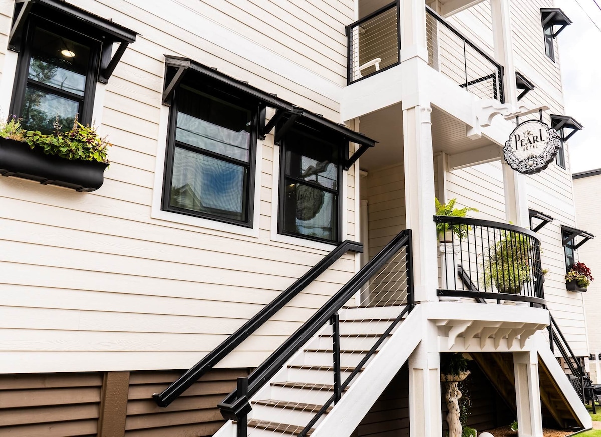 The image shows the exterior of a multi-level building with a light-colored wood facade. A balcony with a black railing is visible, alongside several windows adorned with flower boxes. The entrance is marked by a sign that reads 'Pearl' and a staircase leading up to the entryway.