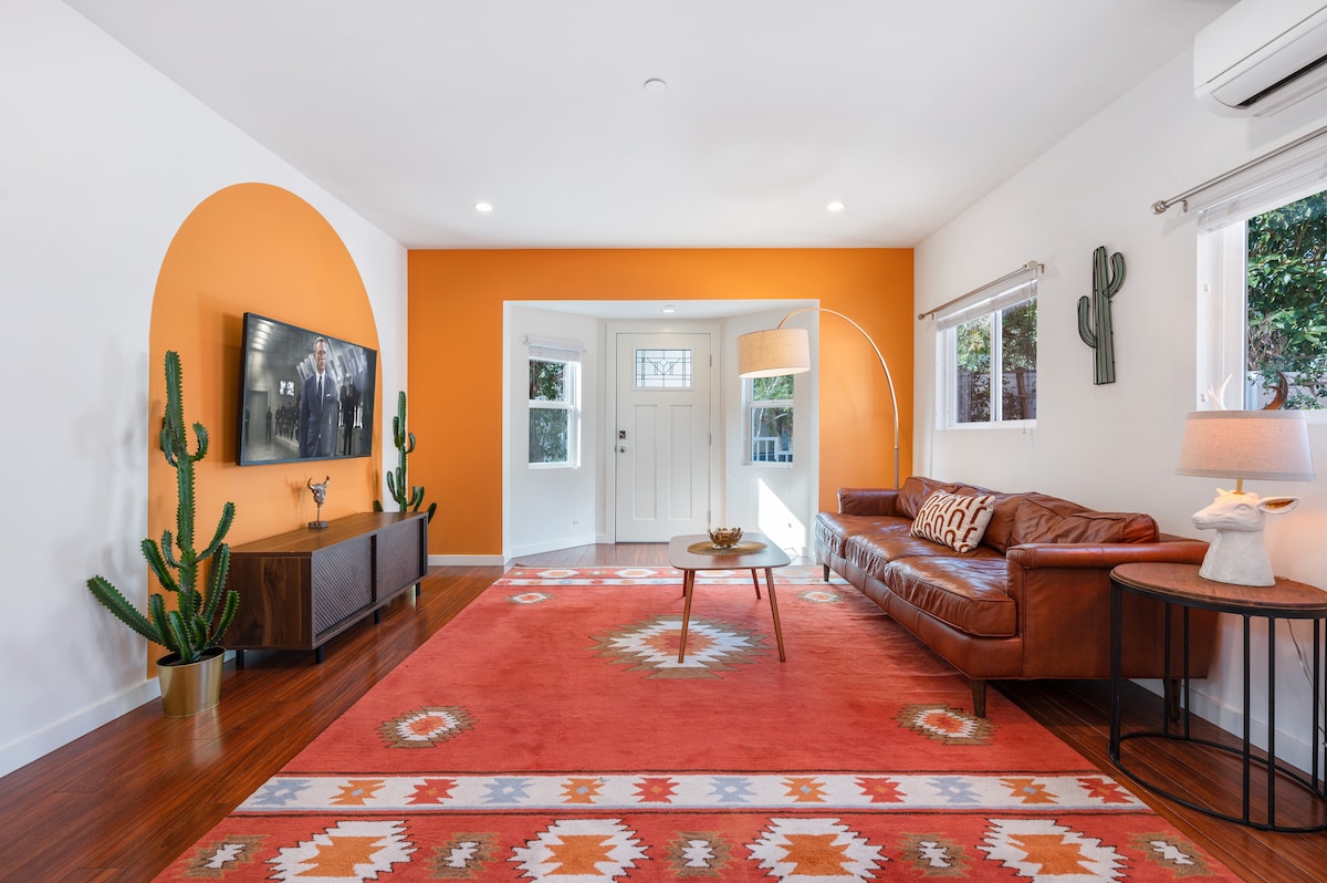 A welcoming living space features a large flat-screen TV mounted on a wall opposite a comfortable brown leather couch. The orange accent wall adds warmth, while decorative cacti and a table lamp enhance the inviting atmosphere. A colorful area rug anchors the seating area.