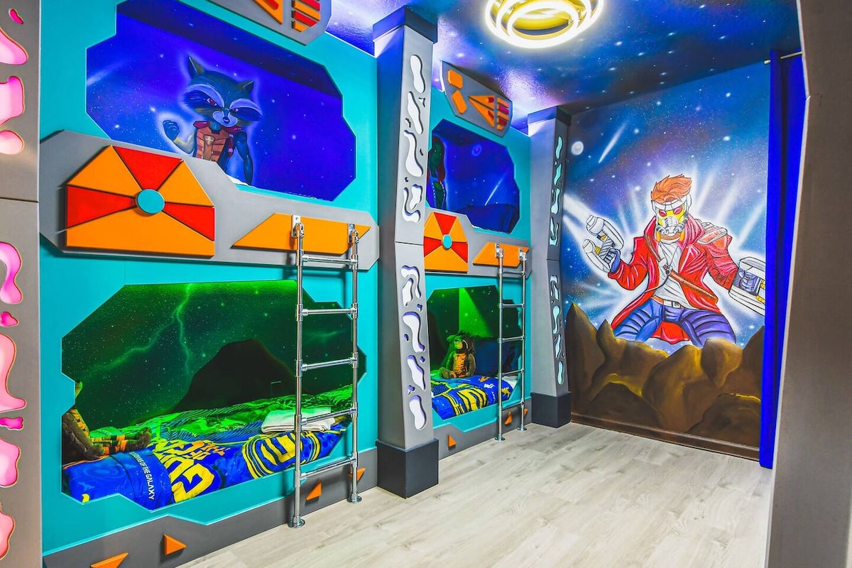 A vibrant Guardians of the Galaxy-themed bedroom features three twin bunk beds with colorful bedding. The walls are decorated with bold graphics, including a character mural, while bright blue and green cosmic scenes enhance the adventurous atmosphere.