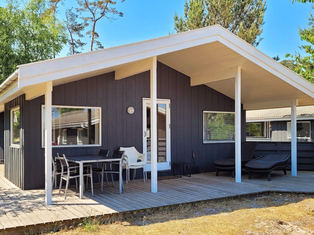A spacious terrace is featured at the entrance of the cottage, with a wooden deck surrounded by trees. Comfortable lounge chairs and a dining table with chairs are visible, offering an inviting outdoor seating area. Large windows provide ample natural light.